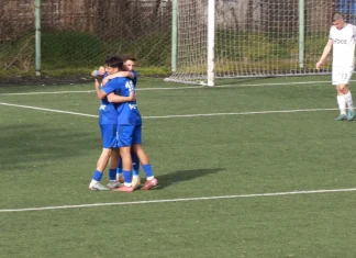 Вторият тим на Левски с убедителен успех у дома Three blue-shirted soccer players hug in celebration on a green turf near the goal net, while a white-clad player walks away in the background.