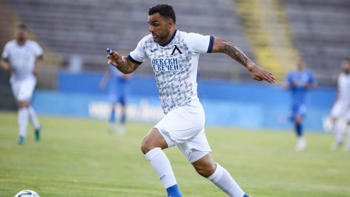 Soccer player in a white and blue geometric jersey runs with the ball on a green pitch, with stadium stands in the background.