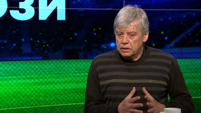 Man with gray hair in a black and gray striped sweater speaks on a TV talk-show set with a green backdrop.