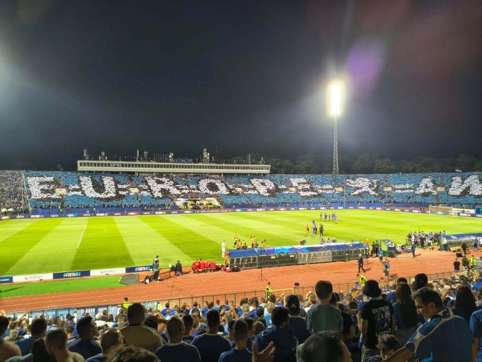 Nighttime soccer match in a large stadium with bright floodlights and a packed blue-and-white banner-filled stands behind the goal.