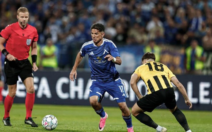 Soccer match: player in blue controls the ball as a defender in yellow and black closes in, with a referee in red on the left and a blurred stadium crowd in the background.