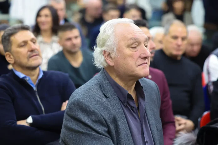 Older man with white hair wearing a gray blazer sits in a crowded audience at a talk or event, looking ahead thoughtfully.