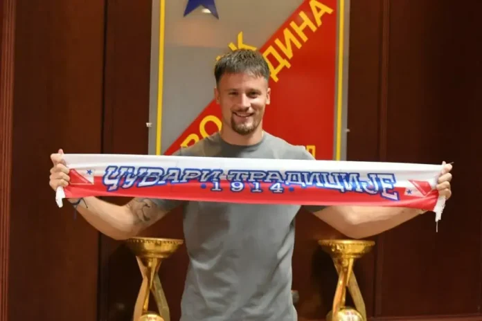 Man in a gray t-shirt holding a long red, white, and blue scarf with Cyrillic text, posing in front of a stage banner.