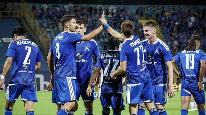 Soccer players in blue jerseys celebrate with high-fives on the field during a match, crowd in the stands behind them.
