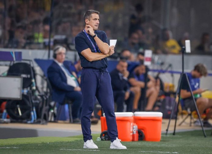 Coach stands on sideline holding a sheet of paper, arms crossed, watching a game on a grassy field.
