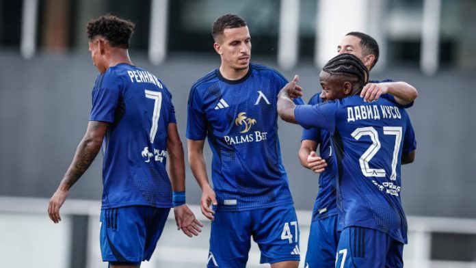 Group of blue soccer players celebrate or train together on the field, one with number 27 on his back, others nearby in mid-conversation.