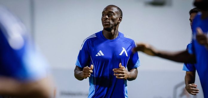 Athlete in a blue training shirt jogs indoors, other players blurred in the background during a drill session.