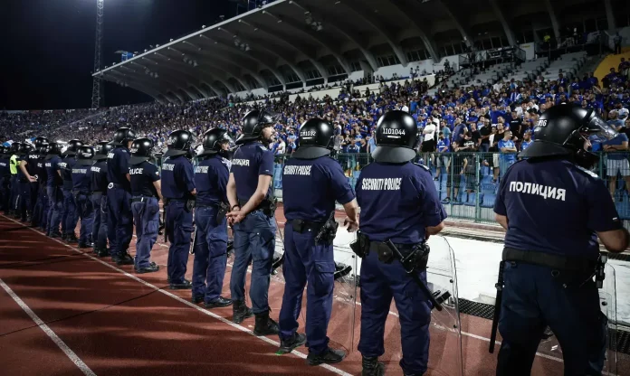 Line of police officers in riot gear stand in a row along a stadium track, facing a large crowd at night stadium.