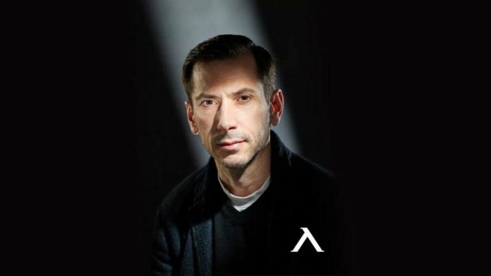 Head-and-shoulders portrait of a man with short dark hair, wearing black clothing, looking at the camera with a neutral expression; white logo resembling an 'A' on his chest against a dark background.