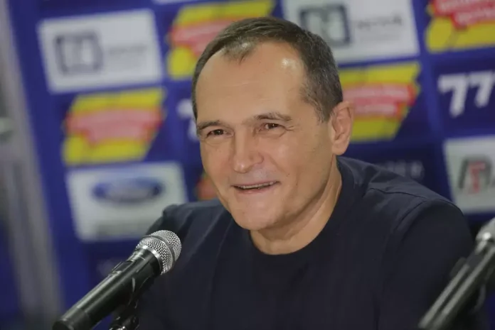 Middle-aged man in a dark shirt speaks into a microphone at a press event, logos blurred in the background.