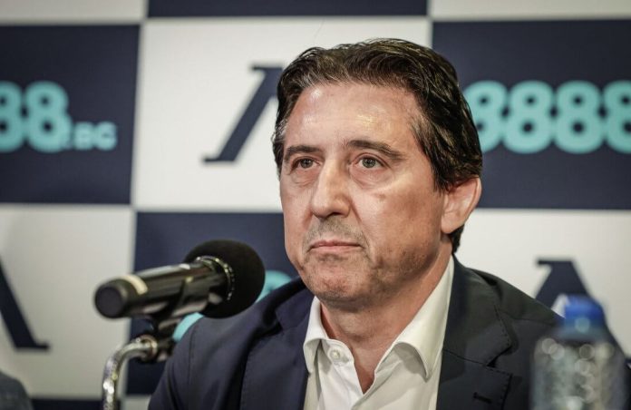 Middle-aged man in a white shirt and dark blazer at a press conference, speaking into a microphone.