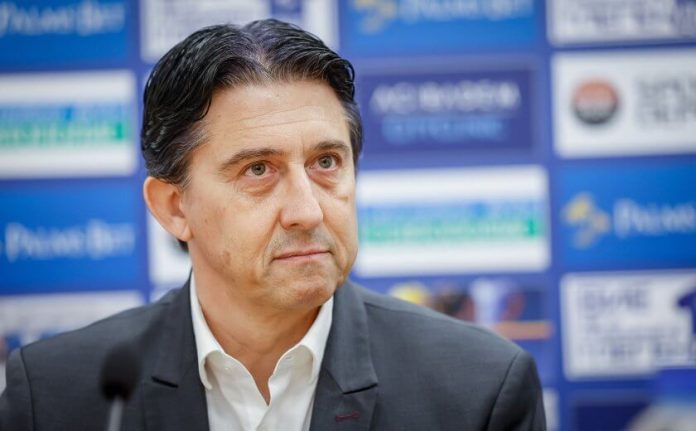 Man in a dark suit and white shirt at a press conference, looking to the side with a blue sponsor backdrop behind him.