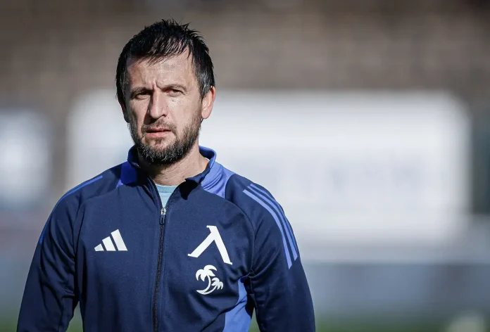 Man in a navy blue Adidas track jacket with white emblem (palm tree) standing outdoors, looking at the camera with a serious expression.