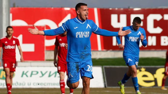 Soccer player in a blue jersey with arms outstretched celebrates on a sunny field, with teammates in red nearby in the background.