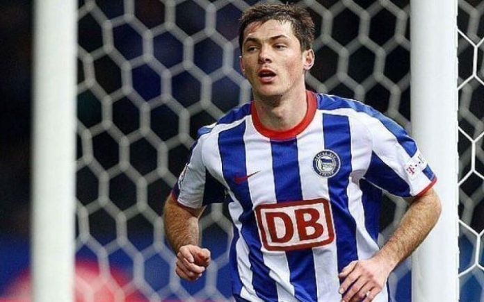 Soccer player in a blue-and-white striped kit runs near the goal net during a match.