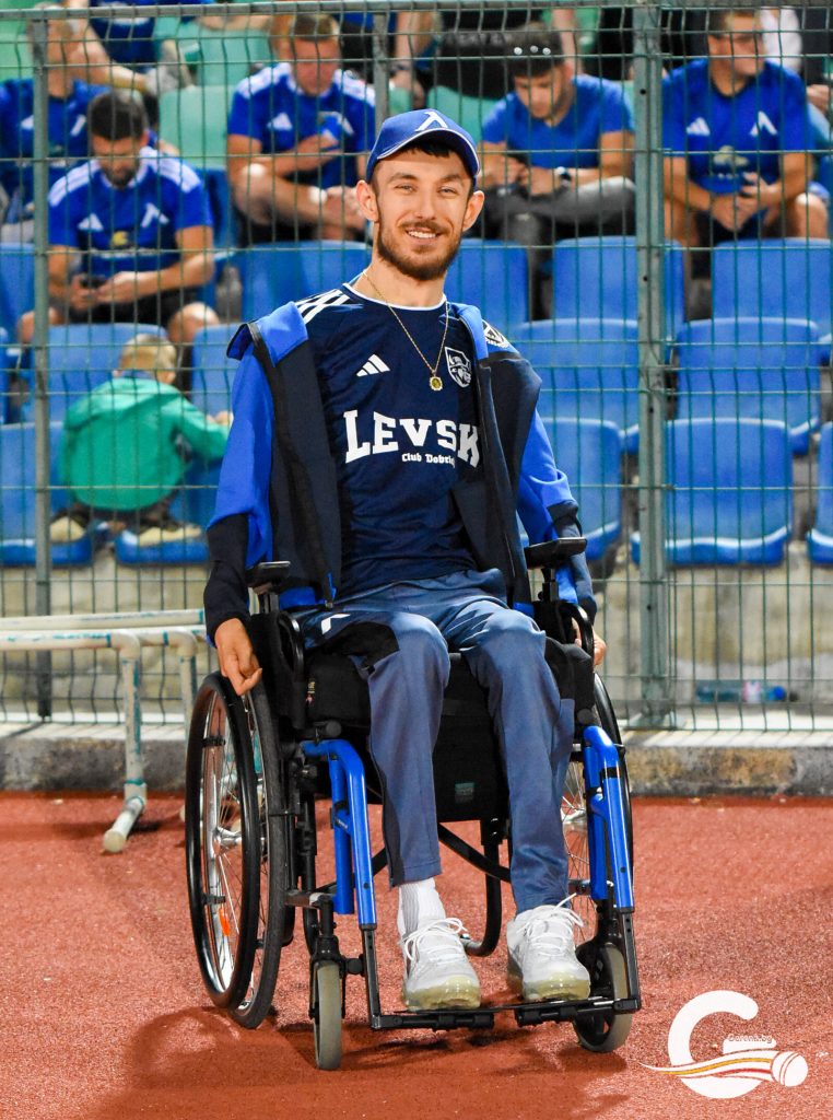 Athlete in a blue sports uniform in a wheelchair on a red track, smiling, with spectators behind a fence in blue shirts.