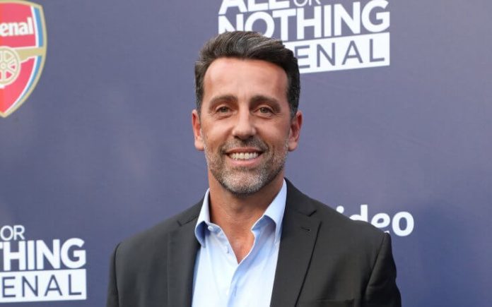 Smiling man in a dark suit and light blue shirt posing in front of a blue backdrop with Arsenal branding and event logos visible.