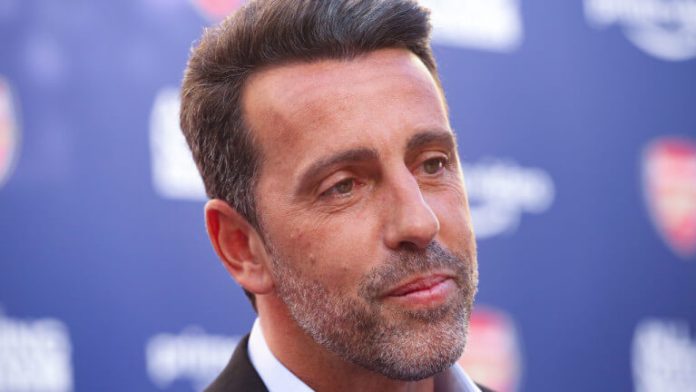 Close-up portrait of a man with salt-and-pepper beard in a suit, facing right, at a blue backdrop event backdrop.