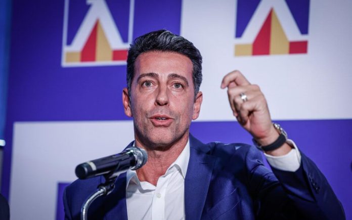 Man in a navy suit speaks into a microphone on stage, raising a hand gesture while addressing an audience.