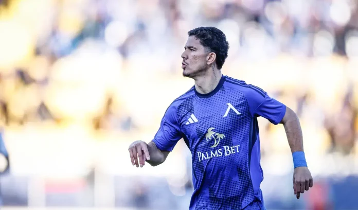 Male soccer player in a blue-purple kit runs on the field, focused and ready to play, Palms Bet sponsor logo visible on his jersey.