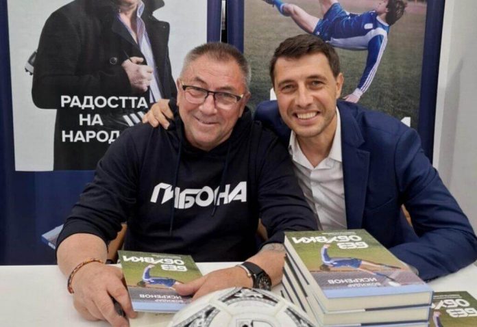 Two men sit at a table signing copies of a book, with stacks of books and a football on the table; one wears a black hoodie with Cyrillic text and both smile at the camera.