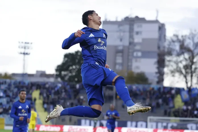 Soccer player in a blue kit leaps mid-air during a match, with stands and opposing players in the background.