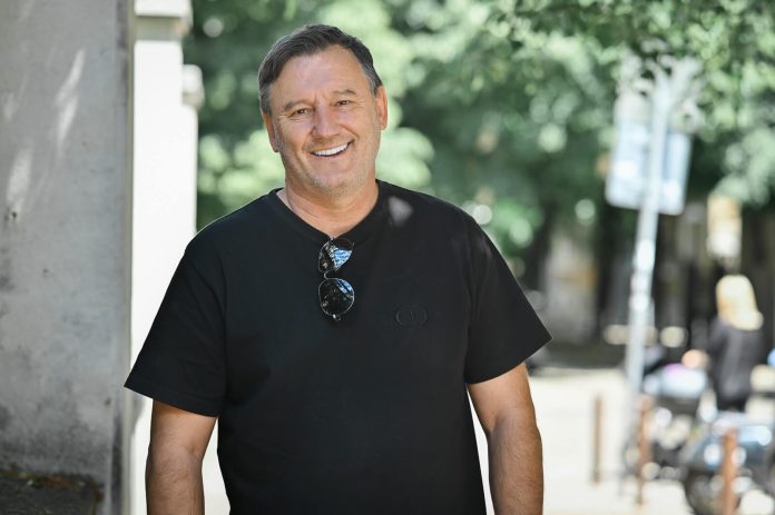 Smiling man in a black t-shirt standing beside a concrete pillar on a sunny street, sunglasses hanging from his neckline.