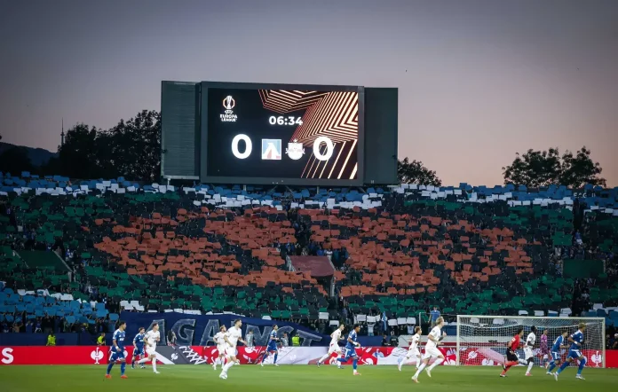 Soccer stadium at dusk with players on the field and a large LED scoreboard showing 0-0 and time elapsed.