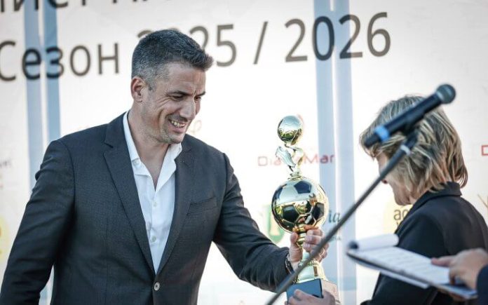 Man in a dark suit smiles as he holds a tall gold trophy on a stage during an award ceremony, a woman with a microphone nearby.