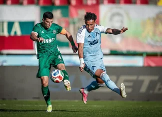 Иван Горанов: Отборната игра ни донесе точката Two soccer players in a green and light-blue kit battle for the ball mid-air on a sunny pitch. The green-clad player wears an orange captain’s armband and kicks toward the ball as the light-blue player closes in.