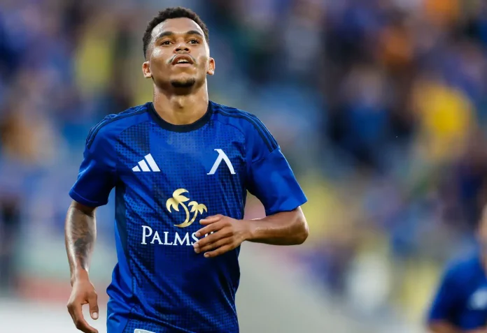 Male soccer player in a blue kit runs on the field during a match, with a palm-tree sponsor logo visible on the chest.