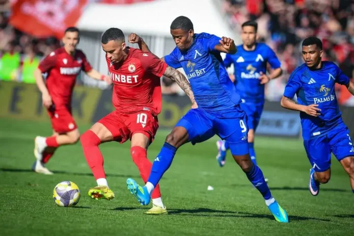 Soccer action: a red-jersey player controls a purple-yellow ball while blue-jersey players chase him on a green field.