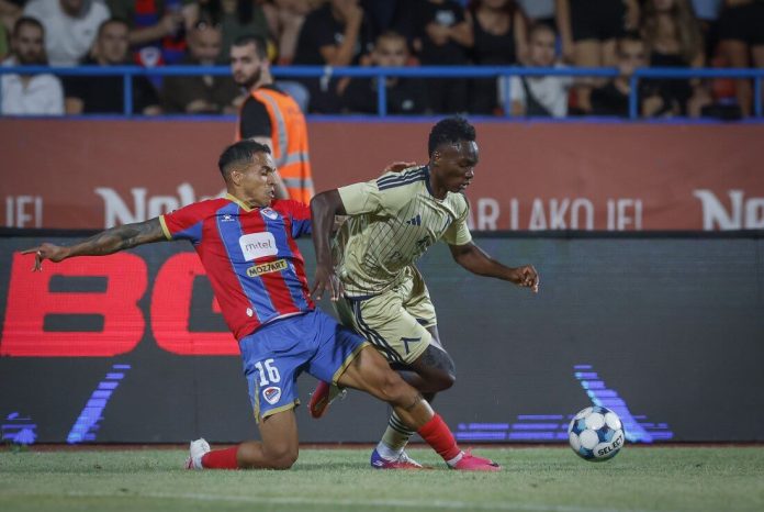 Two soccer players contest the ball on the field, one in red-blue kit and the other in beige, with fans in the stands behind them.