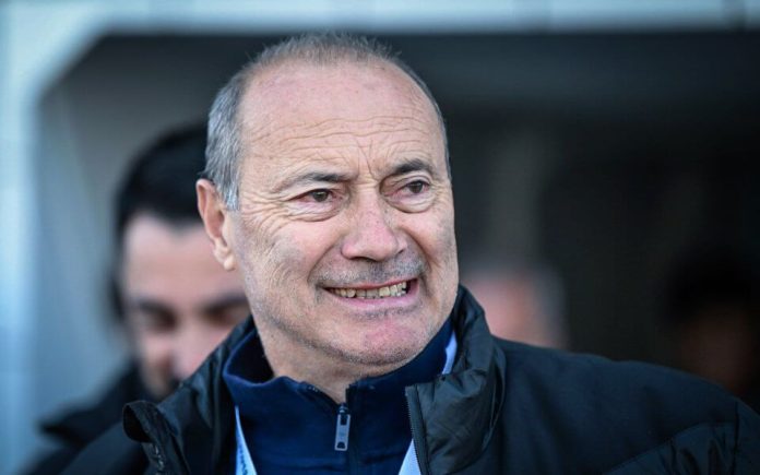 Smiling older man in a dark jacket outdoors, facing slightly to the right, with a blurred background behind him.