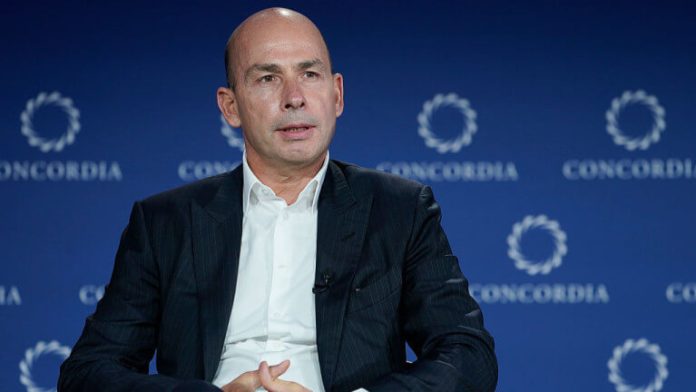 Man in a dark suit and white shirt seated on stage, speaking at a Concordia event with a blue backdrop behind him.