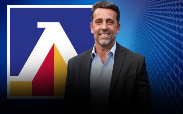 Smiling man in a dark suit stands in front of a blue backdrop with a large white and colored logo behind him.