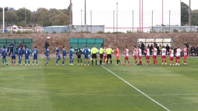 Two soccer teams in blue and red uniforms line up on the field for a pre-match handshake with referees nearby.