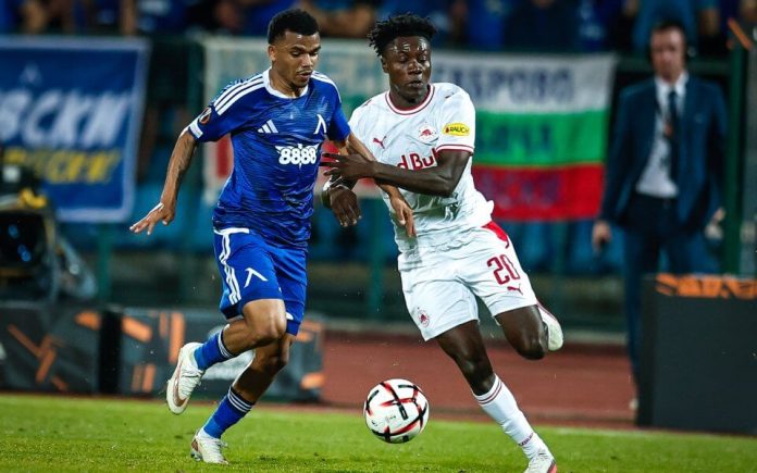 Two soccer players chase a ball during a match, one in blue and one in white, with a crowd in the background.