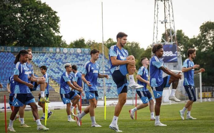 Group of soccer players in blue training kits performing leg raises on a sunny practice field with stadium seating in the background.