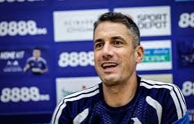 Man in a navy sports shirt speaking at a press conference, with a blue sponsor backdrop behind him.