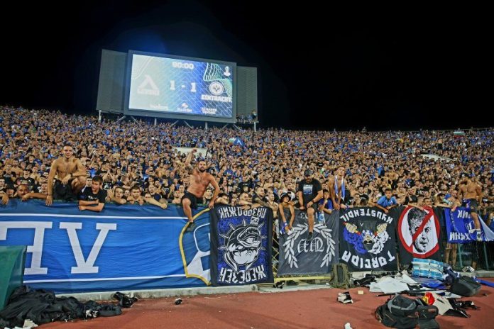 Full stadium crowd in blue, fans leaning over the barrier with banners during a night football match when the scoreboard shows 1-1.