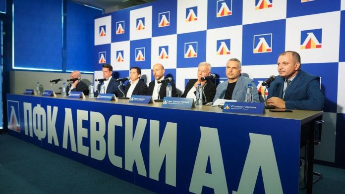 Seven men in suits sit at a long blue table with microphones during a press conference, backdrop covered in logos and blue-white tiles.