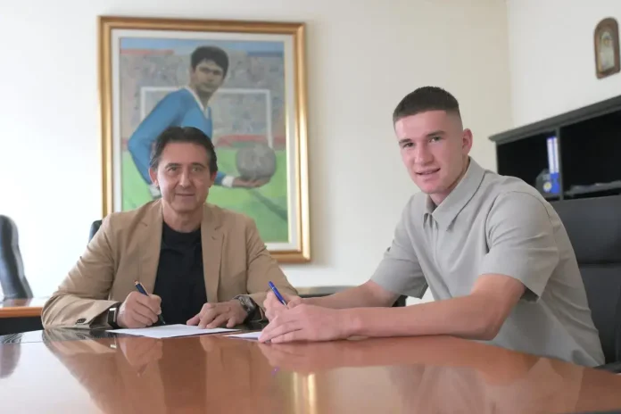 Two men sit at a wooden conference table signing documents in an office, with a large framed painting of a person in blue on the wall behind them.