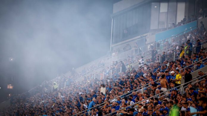 Large crowd of football fans in blue in a stadium, with thick smoke filling the upper stands.