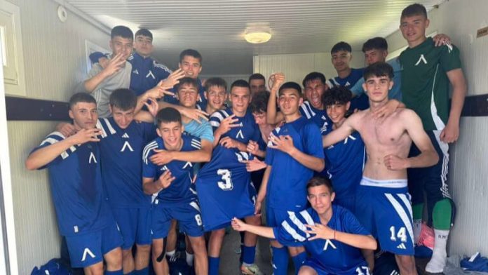 Group of teenage soccer players in blue uniforms posing together in a locker room, some making hand signs, arms around each other in celebration.