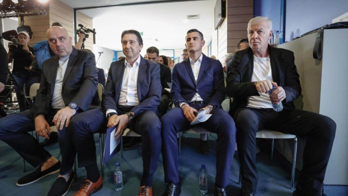 Four men in dark suits sit in a row at a press event, photographers and equipment visible in the background.