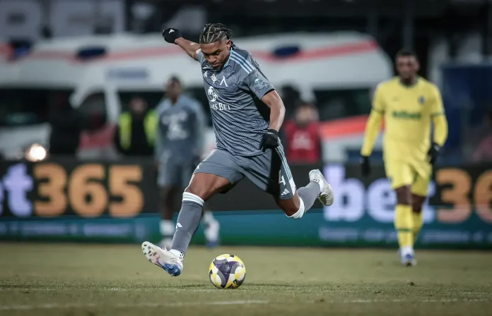 Soccer player in gray kit striking a ball during a match, with a yellow-clad opponent in the background on a grassy field