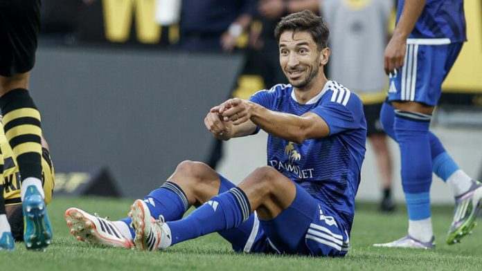 Soccer player in a blue kit sits on the grass during a match, smiling and gesturing with his hands.