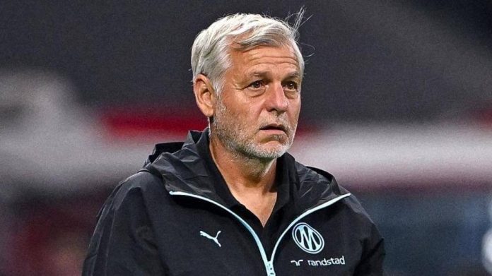 Football coach with gray hair in a black jacket, standing on the sidelines during a match with team logos visible on his chest.