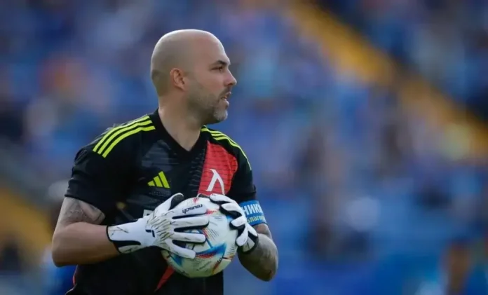 Bald male soccer goalkeeper wearing a black jersey with yellow stripes, holding a ball in white gloves on a field.